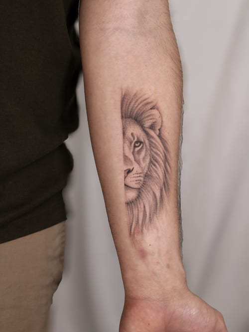 A realistic black and grey lion's face tattoo on the forearm with detailed shading and bold lines — by Melanie Benoit, los angeles