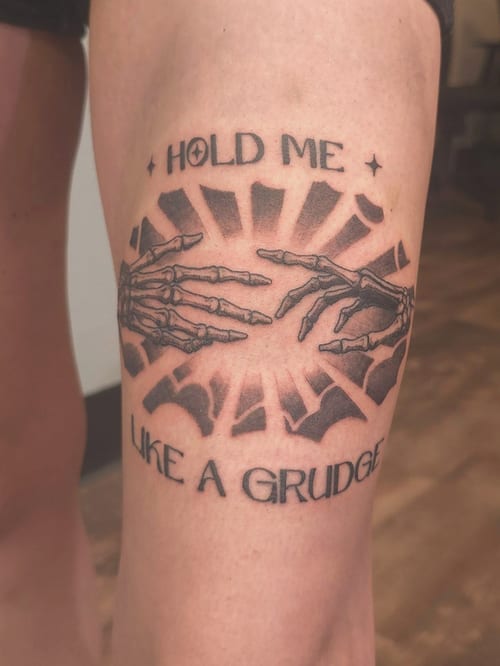 A bold black and grey tattoo of skeleton hands reaching towards each other with the text 'Hold Me Like a Grudge' on the thigh — by Miguel ANGELO, los angeles