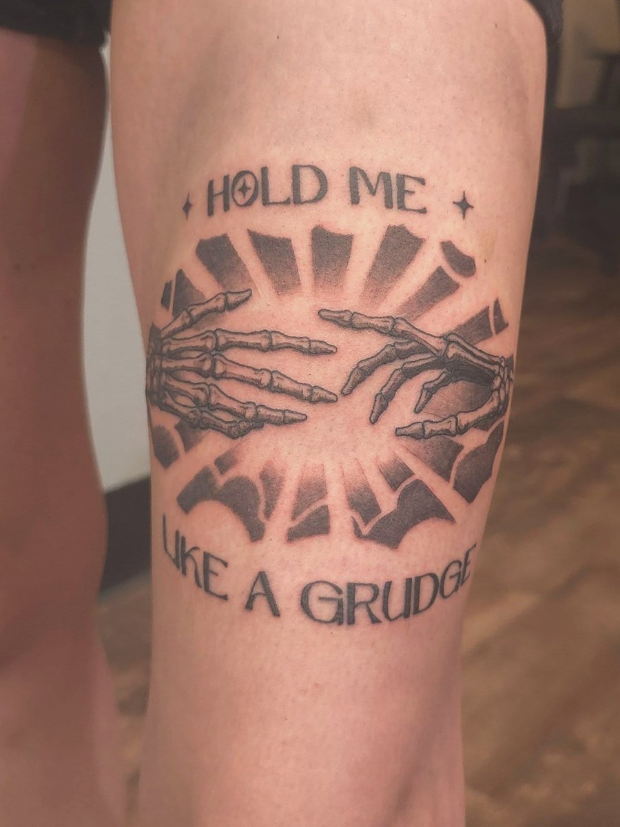 A bold black and grey tattoo of skeleton hands reaching towards each other with the text 'Hold Me Like a Grudge' on the thigh — by Miguel ANGELO, los angeles