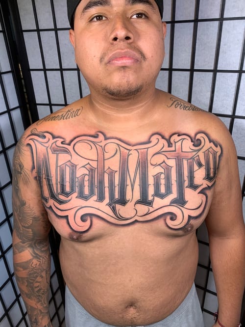 A bold black and grey lettering tattoo across the chest with intricate script and decorative flourishes — by Humilde tattoos, los angeles