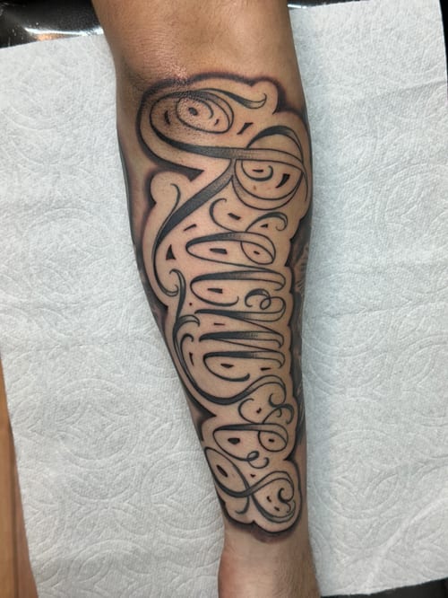 A bold black and grey lettering tattoo on the calf with intricate script design — by Humilde tattoos, los angeles