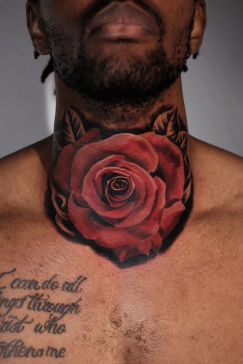 A realistic rose tattoo on the neck with vibrant red petals and black shading, accompanied by script text on the chest — by Johnny Ayala, los angeles