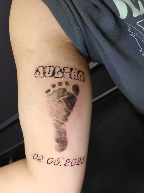 A black and grey tattoo of a baby's footprint with the name 'Julian' and a date on the upper arm, featuring bold lettering — by anoetic.designs.ultd, los angeles
