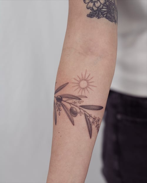 A delicate black and grey fine line tattoo of an olive branch with a sun on the forearm — by Melanie Benoit, los angeles