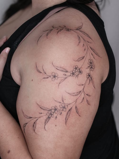 A delicate fine line floral vine tattoo on the shoulder, featuring small flowers and leaves in black and grey — by Melanie Benoit, los angeles