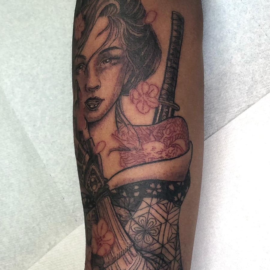A black and grey tattoo of a woman with a sword and flowers on the arm, featuring Japanese style elements — by Sandra Price, los angeles