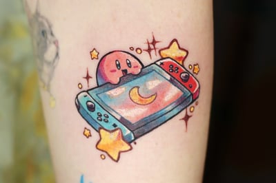 A colorful tattoo of Kirby with a gaming console, surrounded by stars and a crescent moon, in a playful sticker style on the arm — by Tweetwee, los angeles