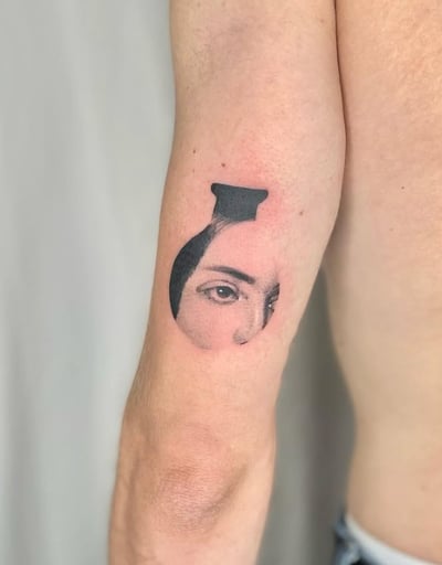 A black and grey abstract tattoo on the upper arm featuring a partial face with a focus on the eyes, enclosed in a circular shape — by Level tattoo, los angeles