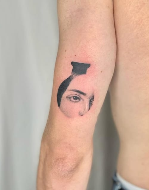 A black and grey abstract tattoo on the upper arm featuring a partial face with a focus on the eyes, enclosed in a circular shape — by Level tattoo, los angeles