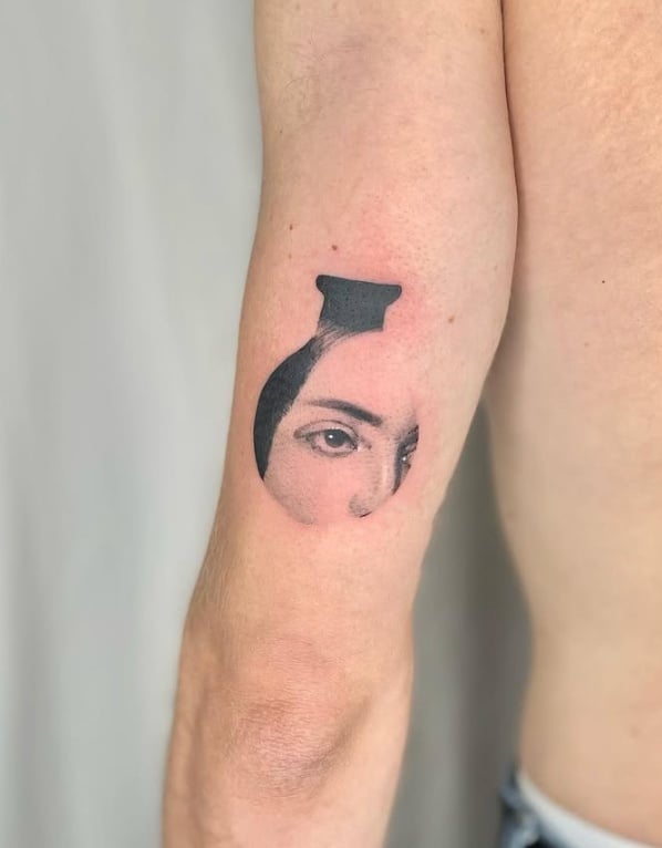 A black and grey abstract tattoo on the upper arm featuring a partial face with a focus on the eyes, enclosed in a circular shape — by Level tattoo, los angeles