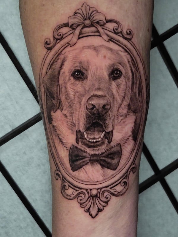 A realistic black and grey dog portrait tattoo on the forearm, featuring ornate framing and a bow tie — by Miguel ANGELO, temecula, ca