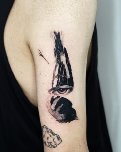A black and grey surrealistic tattoo on the upper arm featuring a bird integrated with a human eye, creating a striking visual effect — by Level tattoo, los angeles