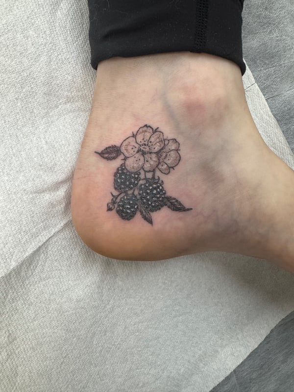 A delicate black and grey tattoo of a blackberry plant with flowers on the ankle, featuring a minimal and botanical style — by Sandra Price, ventura, ca