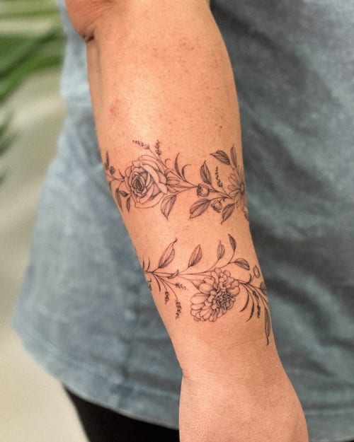 A delicate black and grey floral band tattoo on the forearm featuring roses and other flowers in fine line style — by Melanie Benoit, los angeles