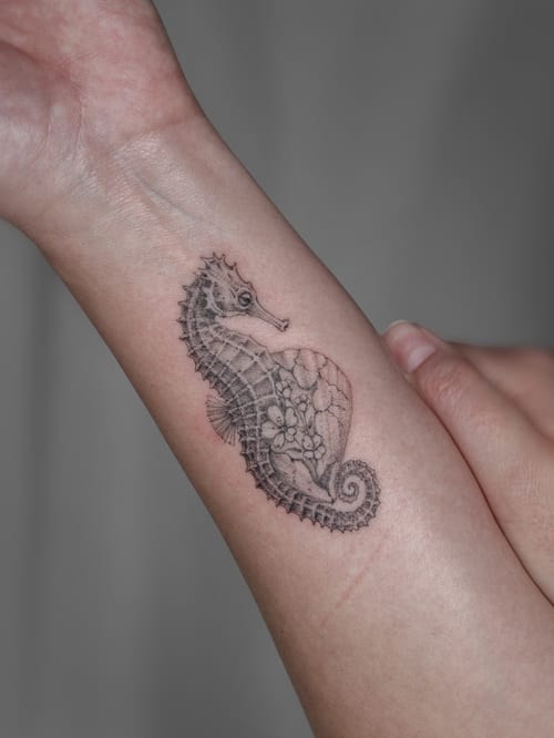 A delicate black and grey seahorse with floral design on the forearm, featuring fine line and blackwork style — by Melanie Benoit, los angeles