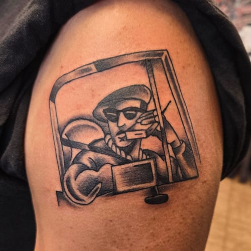 A black and grey illustrative tattoo of a person in a car, wearing sunglasses and holding a radio, located on the upper arm — by Dretty, los angeles