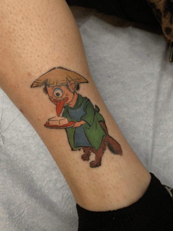 A colorful illustrative tattoo of a mythological creature with one eye, wearing a hat and holding a plate, located on the calf — by tyler_whitlock, los angeles