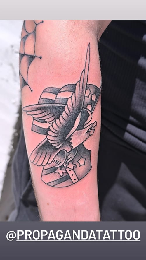 A bold black and grey eagle with an American flag on the forearm in American Traditional style — by Samuel Connelly, los angeles