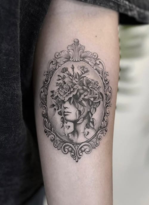A black and grey tattoo on the forearm featuring a woman's face adorned with flowers, framed in an ornamental design with realistic shading — by Jian Liang, los angeles