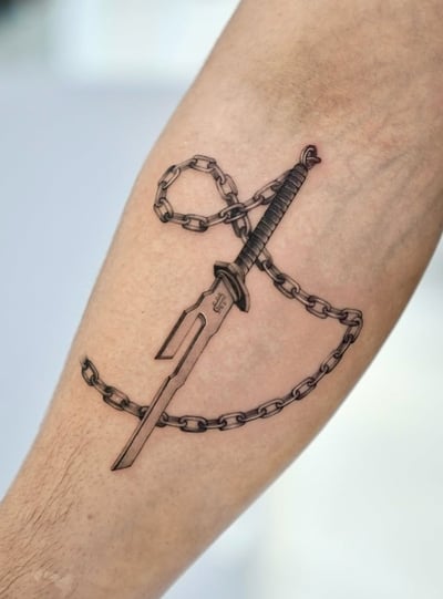 A black and grey realism tattoo of a dagger intertwined with a chain on the forearm, featuring detailed shading and texture — by otaybeatdown, los angeles