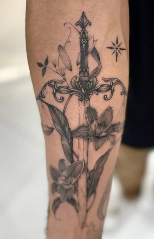 A black and grey illustrative tattoo of a sword intertwined with flowers on the forearm, featuring fine line details and elegant shading — by otaybeatdown, los angeles