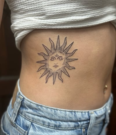 A minimalist linework tattoo of a sun with a face on the rib, featuring simple black and grey lines — by Sandra Price, los angeles