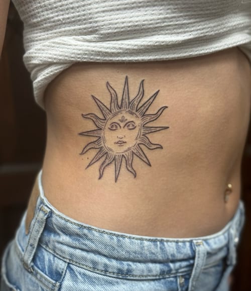 A minimalist linework tattoo of a sun with a face on the rib, featuring simple black and grey lines — by Sandra Price, los angeles