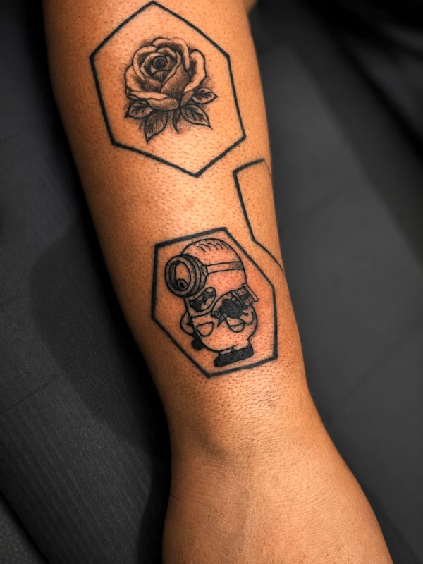 A bold blackwork tattoo featuring a rose and a cartoon character on the forearm, with strong outlines and graphic style — by HEAVY INK, chino, ca