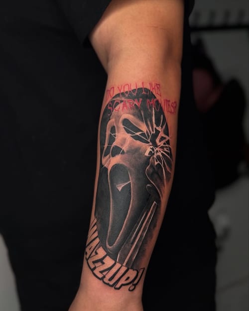 A black and grey tattoo of the Ghostface mask from the Scream movies on the forearm, featuring bold script and realistic shading — by zealous, los angeles