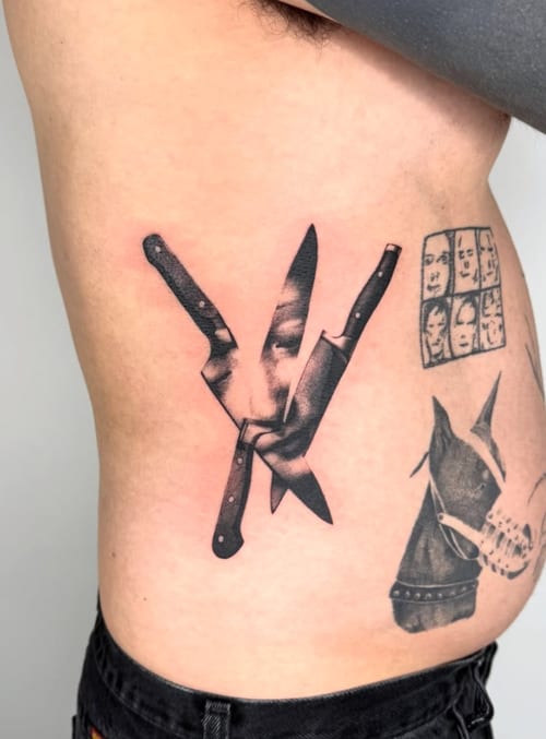 A surreal black and grey tattoo on the rib featuring two knives intersecting with a face partially visible between them, creating a striking and abstract composition — by Level tattoo, los angeles