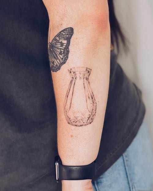 A minimalist black and grey tattoo of a butterfly and a vase on the forearm, featuring fine line work and delicate details — by Level tattoo, los angeles
