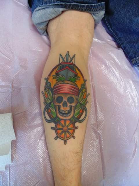 A colorful traditional tattoo of a skull with a bandana and ship wheel on the calf — by Samuel Connelly, san diego, ca