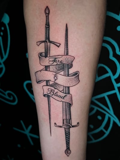 A black and grey tattoo on the forearm featuring two swords with a banner wrapped around them, displaying the words 'Fire & Blood' — by Josue Bravo, los angeles