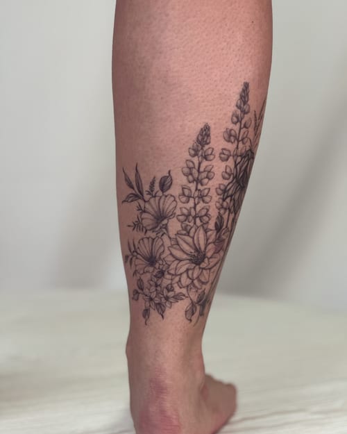 A delicate black and grey floral arrangement tattoo on the calf, featuring fine line work and intricate details — by Melanie Benoit, los angeles