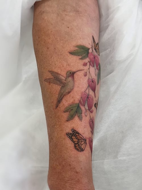 A colorful and delicate tattoo of a hummingbird with flowers and butterflies on the forearm, done in a realistic style — by Melanie Benoit, los angeles