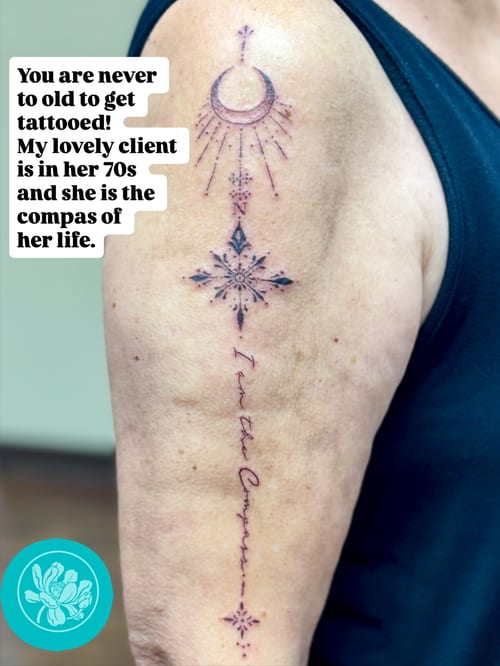 A delicate fine line tattoo on the upper arm featuring a compass and crescent moon with script lettering — by Sandra Price, los angeles