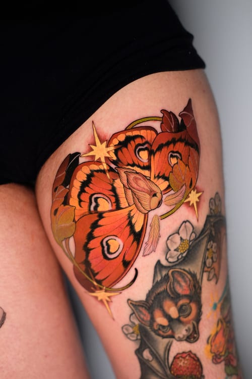 A vibrant Neo-Traditional tattoo on the thigh featuring a large butterfly with intricate patterns, a bat, and flowers, all in rich colors — by Gabe Motta, los angeles