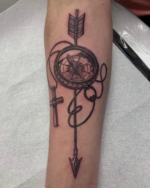 A bold black and grey compass with an arrow and cross on the forearm, featuring detailed linework and graphic elements — by Sandra Price, los angeles