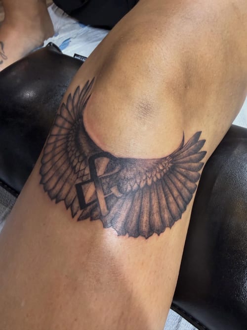 A bold black and grey tattoo of wings with a cross on the thigh, featuring realistic feather details — by Kiera Garvin, los angeles