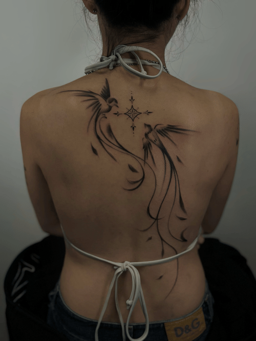 A black and grey ornamental tattoo on the back featuring two stylized birds with flowing lines and floral elements, creating an elegant and symmetrical design — by Rae kaku, los angeles