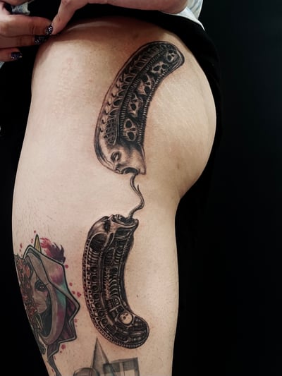 A surreal black and grey tattoo on the thigh featuring two abstract faces with intricate skull patterns, connected by a flowing line — by zitronxus, los angeles