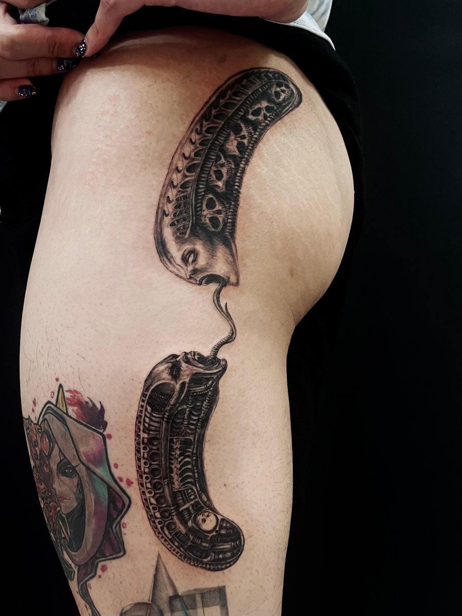 A surreal black and grey tattoo on the thigh featuring two abstract faces with intricate skull patterns, connected by a flowing line — by zitronxus, los angeles