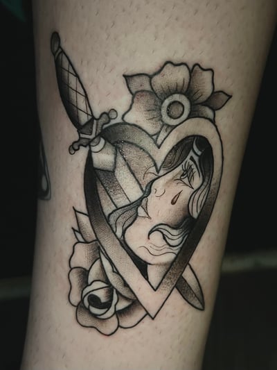 A black and grey Neo-Traditional tattoo on the leg, featuring a heart with a woman's face inside, surrounded by flowers and a dagger piercing through — by Josue Bravo, los angeles