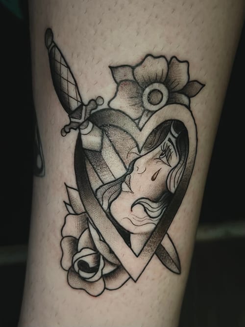 A black and grey Neo-Traditional tattoo on the leg, featuring a heart with a woman's face inside, surrounded by flowers and a dagger piercing through — by Josue Bravo, los angeles