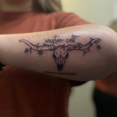 A graphic black and grey tattoo of a bull skull with flowers on the arm — by Maddie Ramirez, los angeles