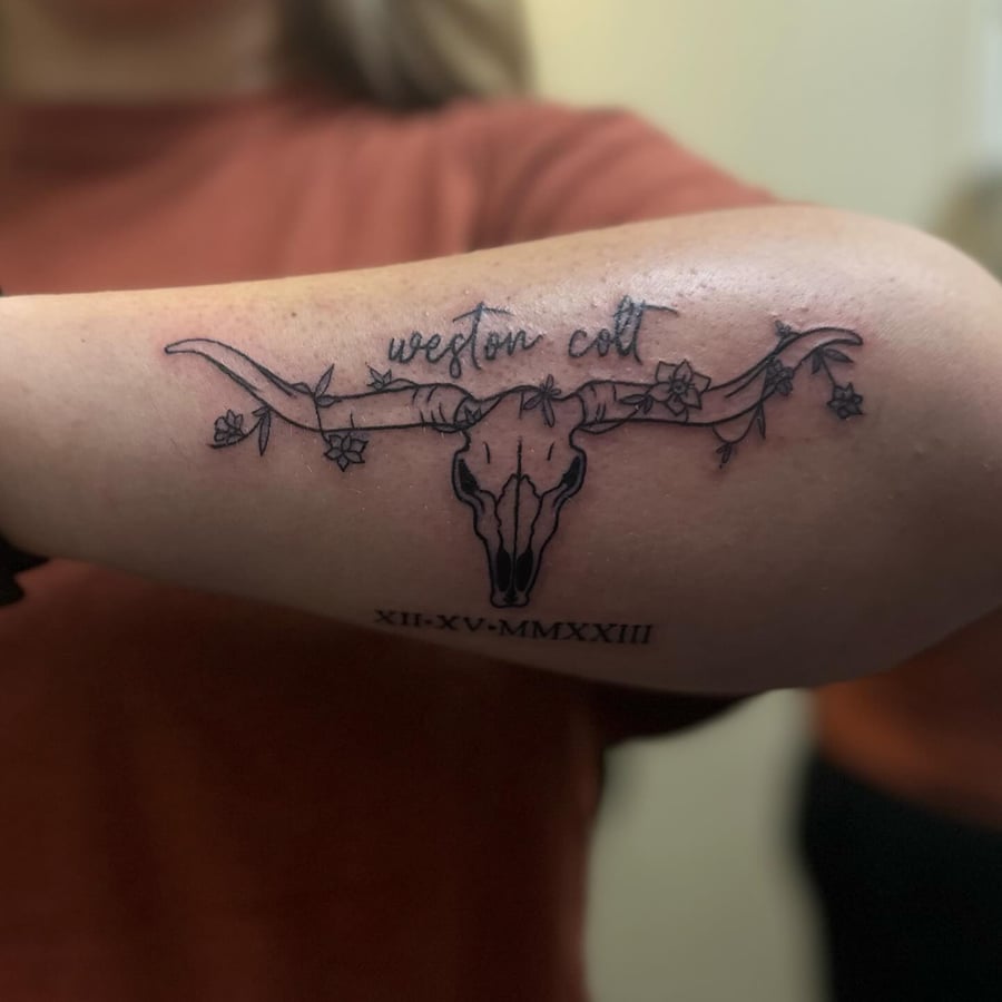 A graphic black and grey tattoo of a bull skull with flowers on the arm — by Maddie Ramirez, los angeles