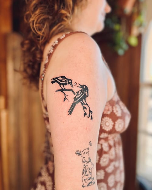 A minimalist blackwork tattoo of two birds on a branch on the upper arm, featuring bold lines and a simple design — by Dylan Aiello, los angeles