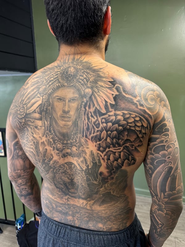 A detailed black and grey realism tattoo covering the full back, featuring a portrait of a man with a headdress and mythological elements — by Humilde tattoos, los angeles
