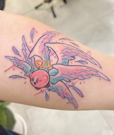 A colorful watercolor tattoo of Kirby with angel wings and a halo on the forearm, featuring vibrant pink and blue hues — by Tweetwee, los angeles