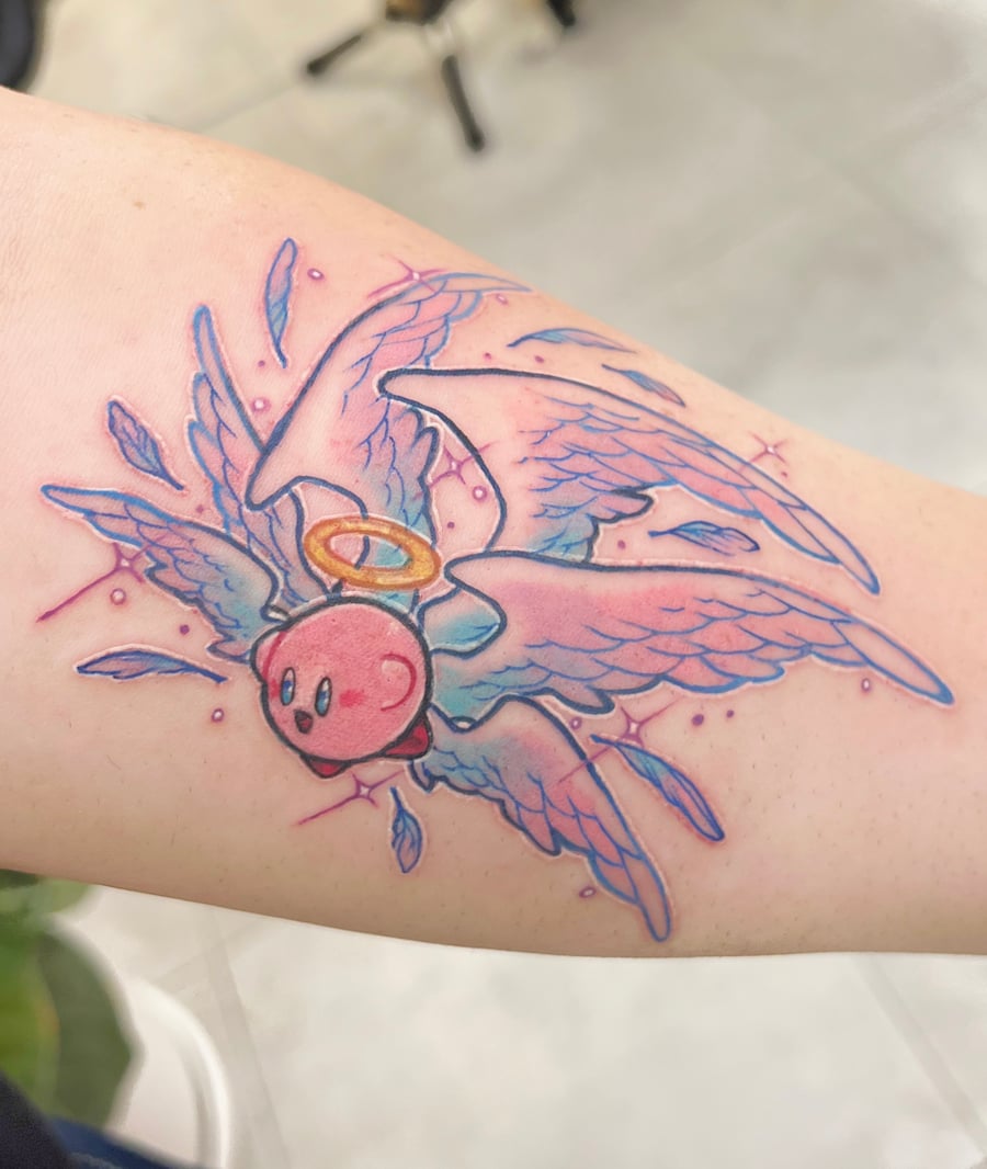 A colorful watercolor tattoo of Kirby with angel wings and a halo on the forearm, featuring vibrant pink and blue hues — by Tweetwee, los angeles